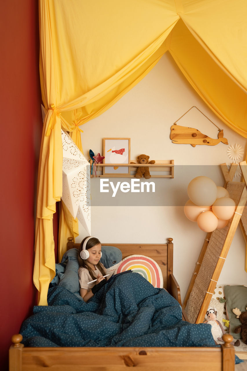 Cute concentrated little girl with long hair in pajama and headphones using stylus while drawing on graphic tablet and sitting on comfortable bed under warm blanket at home