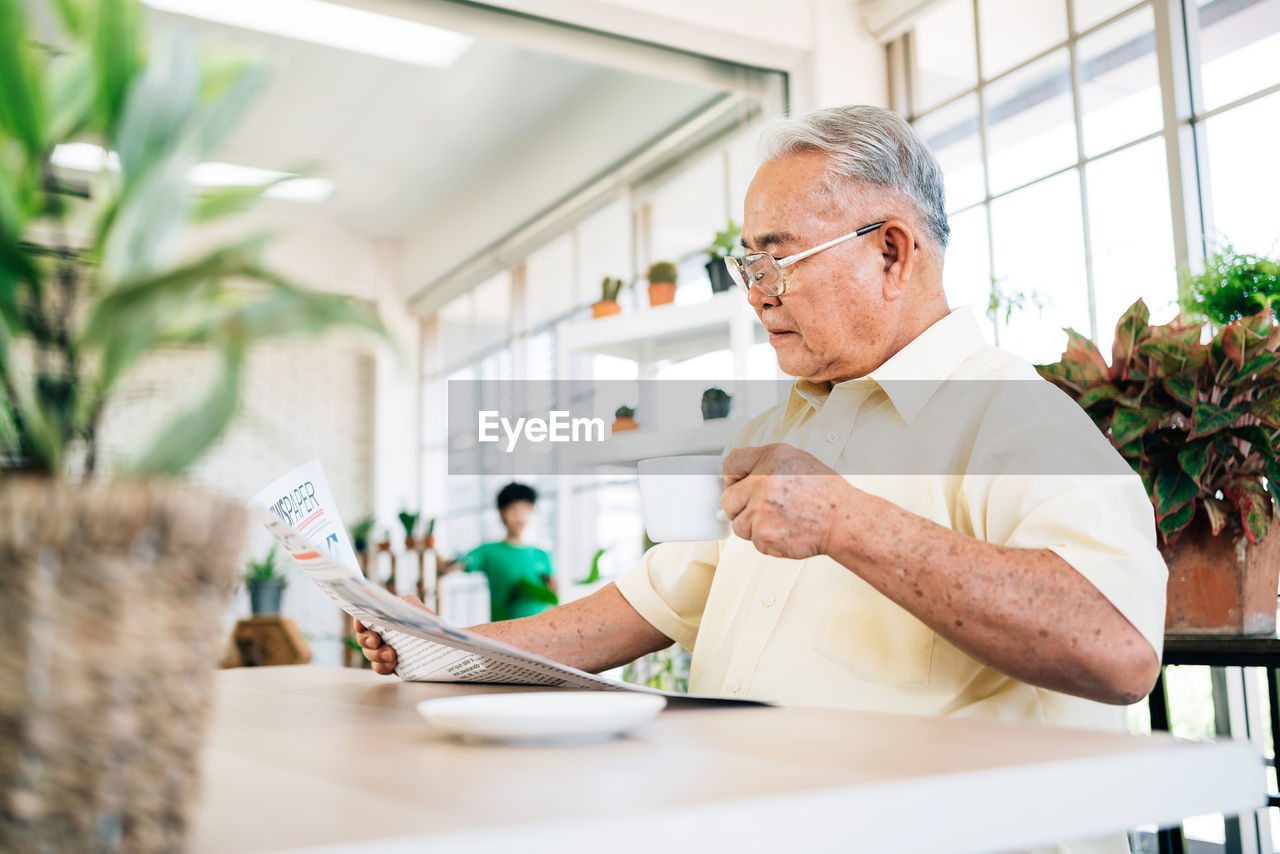 Senior man holding coffee cup while reading newspaper at greenhouse
