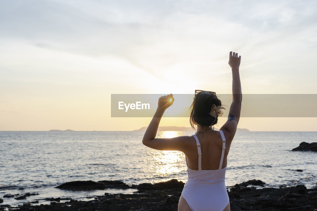 Rear view of woman with arms raised standing at beach against sky during sunset