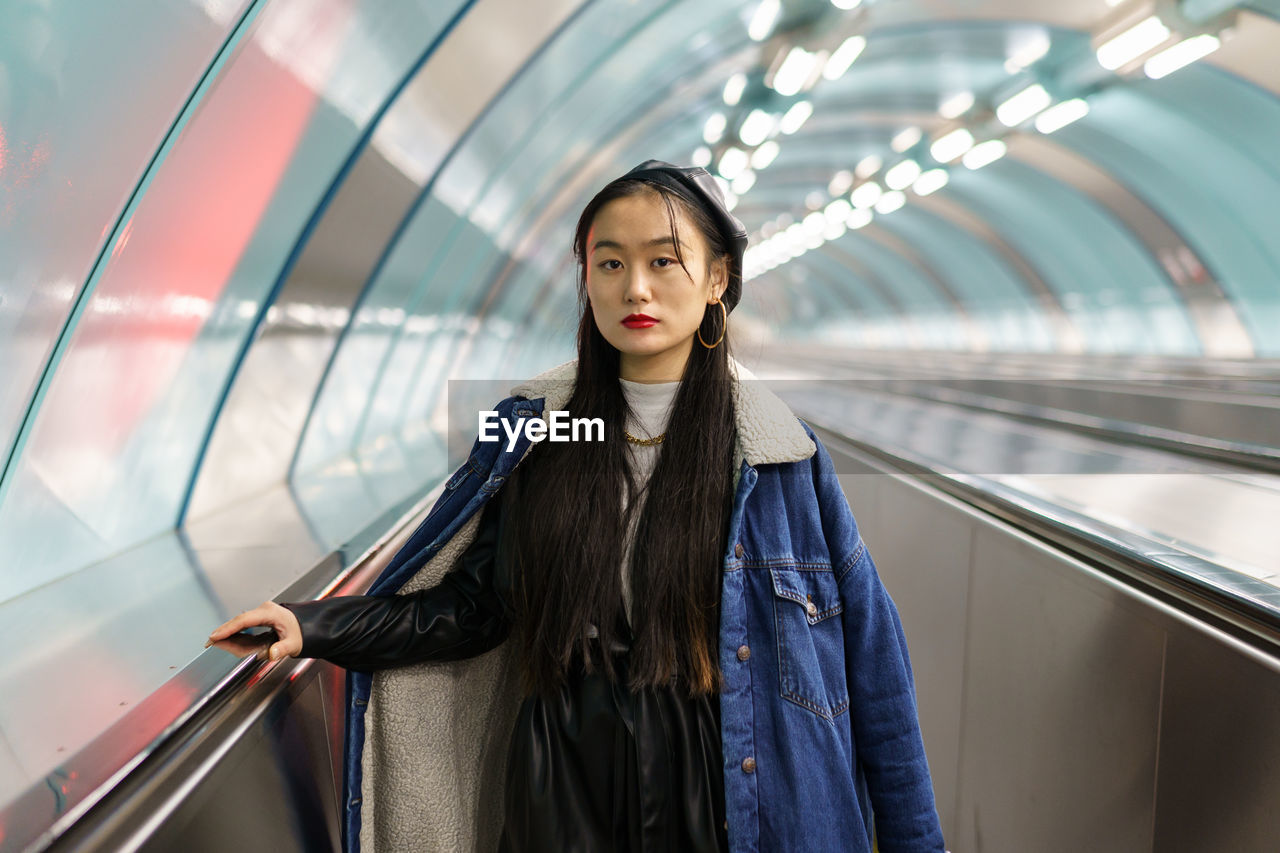 Stylish chinese girl in trendy street fashion outfit, jeans coat, leather beret on subway escalator