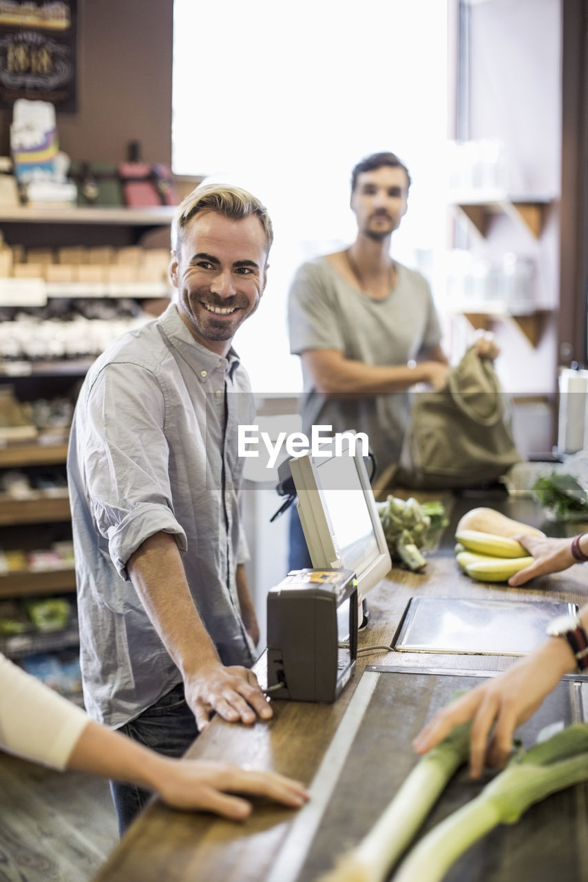 Smiling man standing at supermarket checkout counter with friend in background