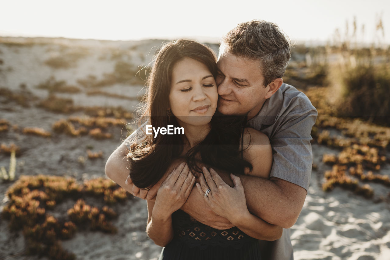 Mid-40's husband embraces beautiful wife with sand dune in background