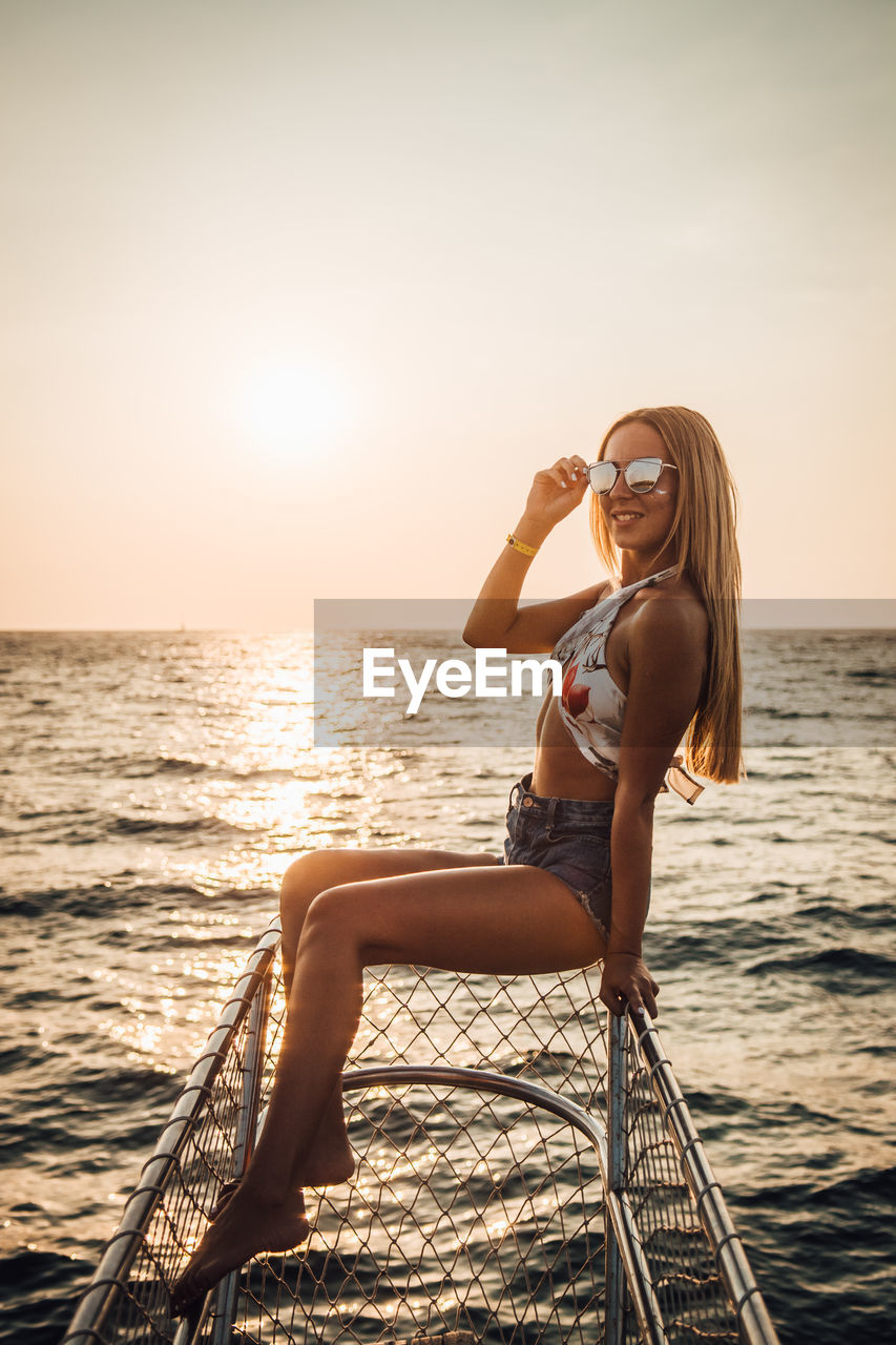 Fashionable young woman sitting on fence by sea against clear sky during sunset