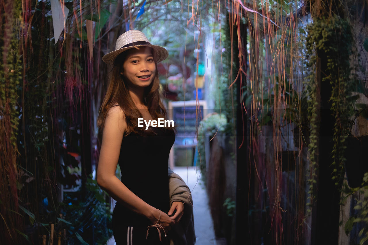 Portrait of smiling woman wearing hat standing amidst creeper plant