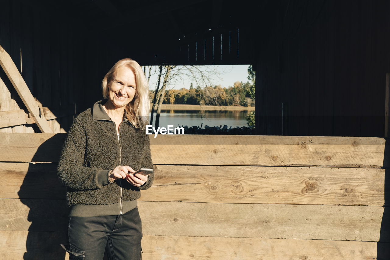 Portrait of smiling mature female farmer using mobile phone outside barn