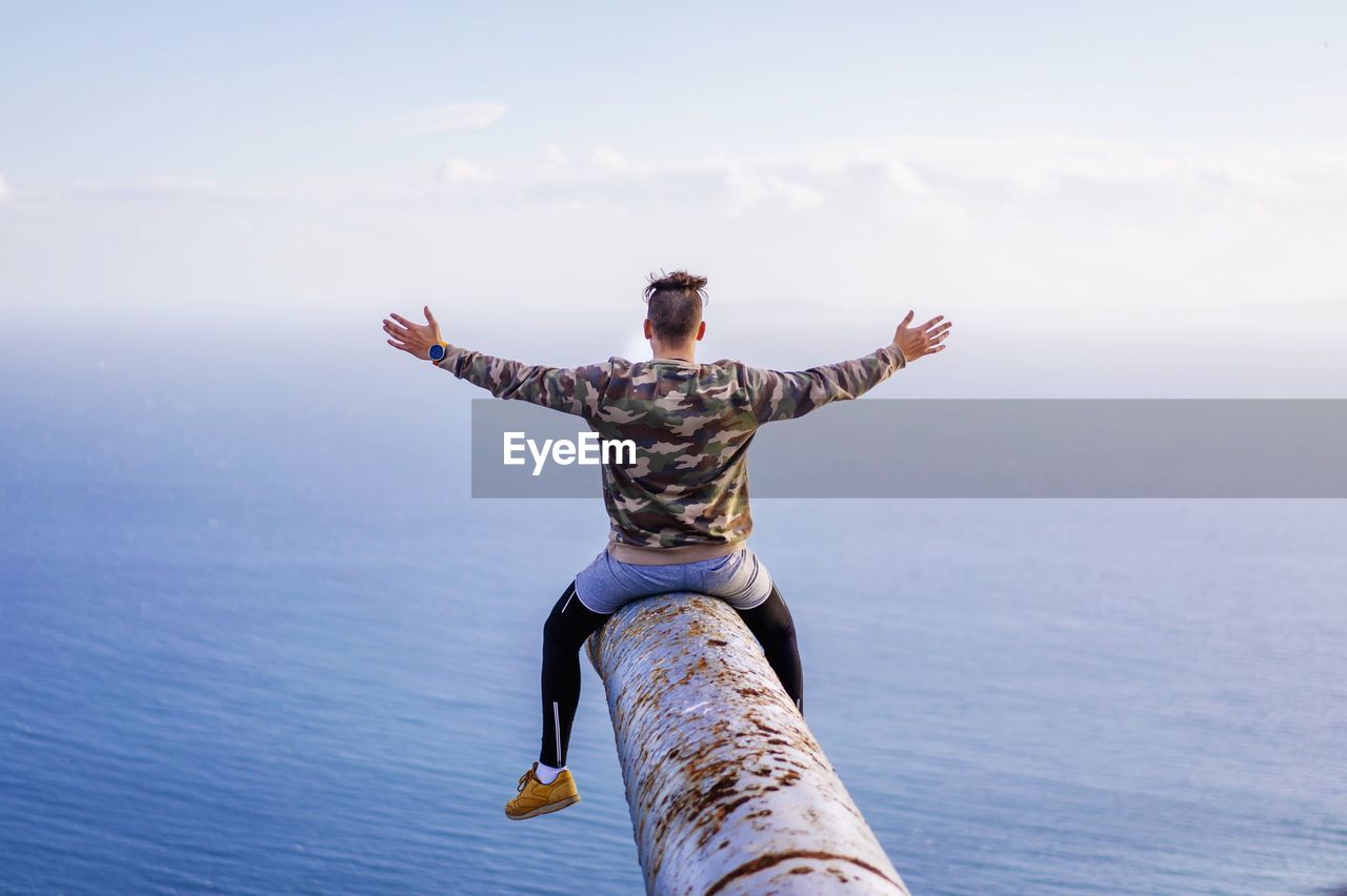 Rear view of man sitting with arms outstretched on rusty metallic pipe while looking at sea against sky