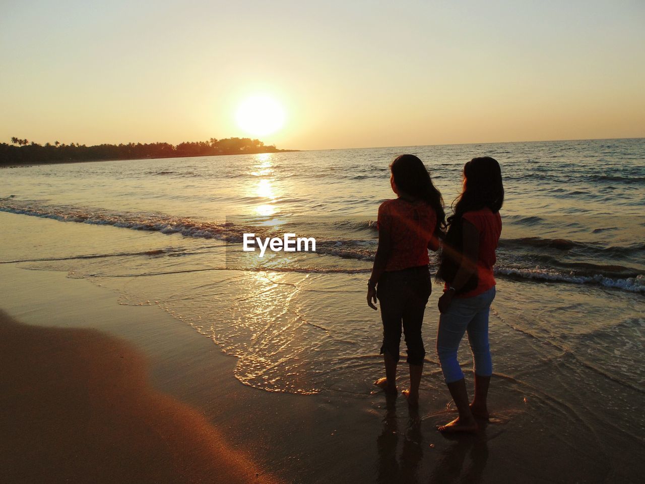 REAR VIEW OF COUPLE STANDING ON BEACH DURING SUNSET