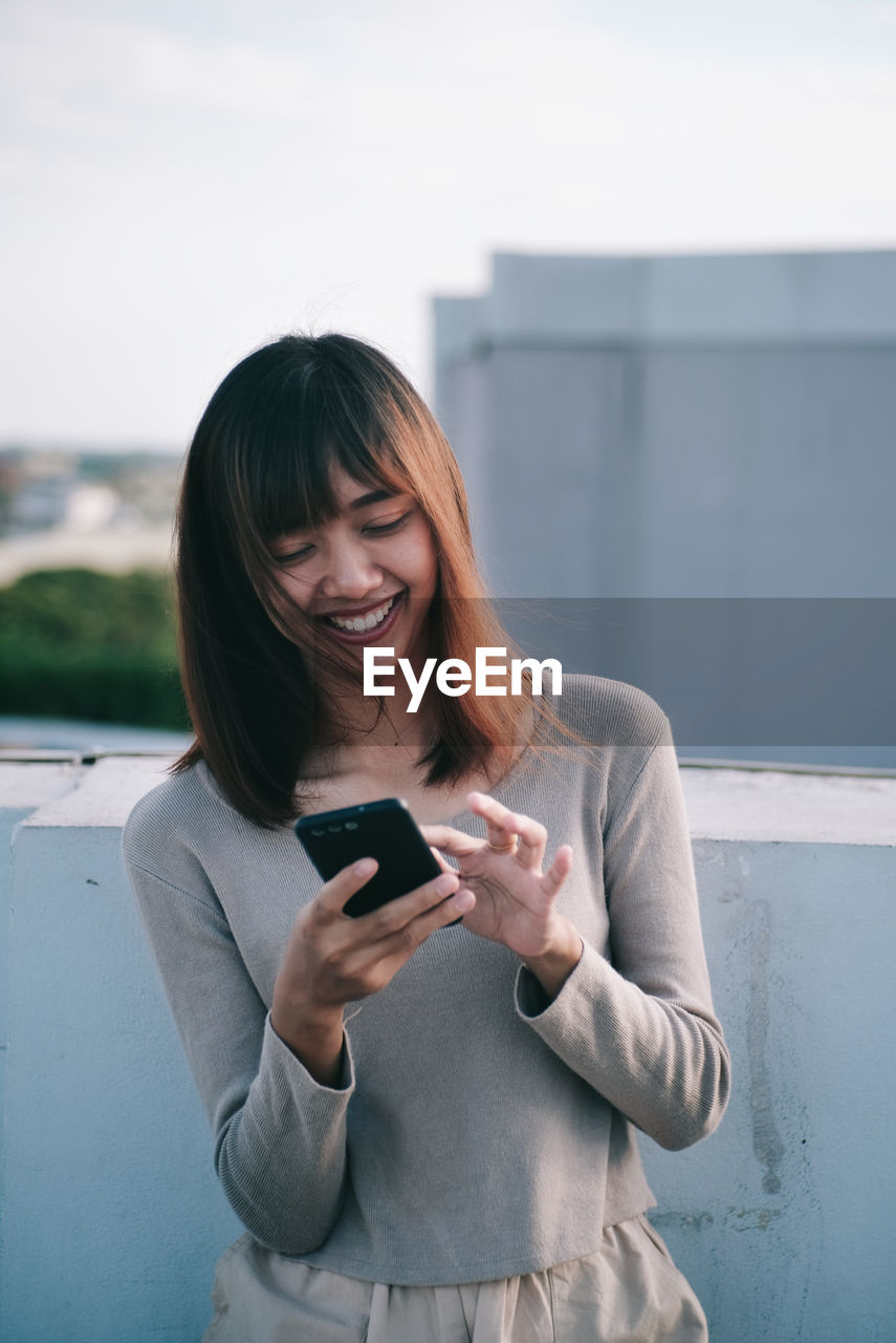 YOUNG WOMAN USING SMART PHONE WHILE STANDING ON MOBILE