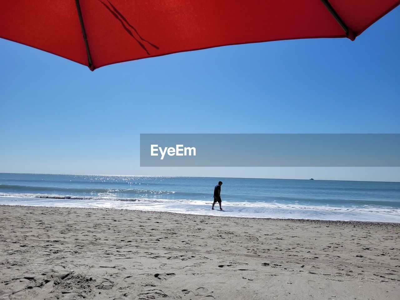 beach, water, sea, land, umbrella, sky, horizon over water, horizon, nature, sand, one person, beauty in nature, clear sky, holiday, ocean, leisure activity, wind, vacation, scenics - nature, trip, adult, day, full length, lifestyles, motion, blue, body of water, tranquility, parasol, sunny, sports, protection, summer, coast, standing, outdoors, tranquil scene, sunlight, relaxation, red, travel destinations, walking, men, environment, wave, women