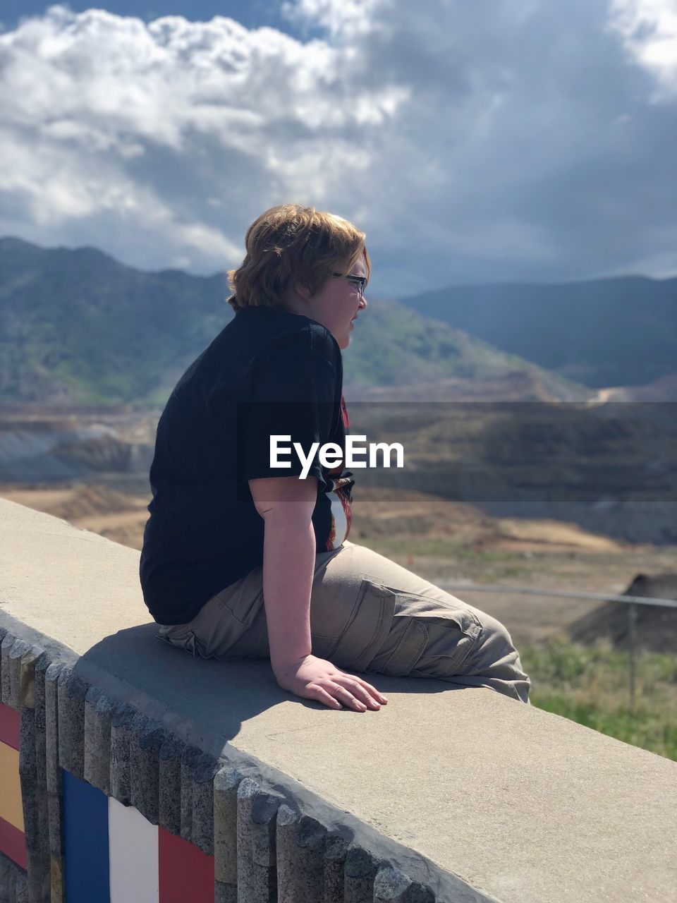 Side view of overweight boy looking at landscape while sitting on retaining wall against sky