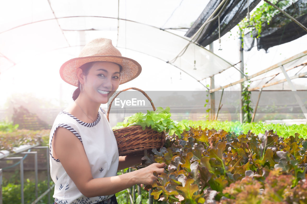 Portrait of happy woman holding vegetables while standing in greenhouse