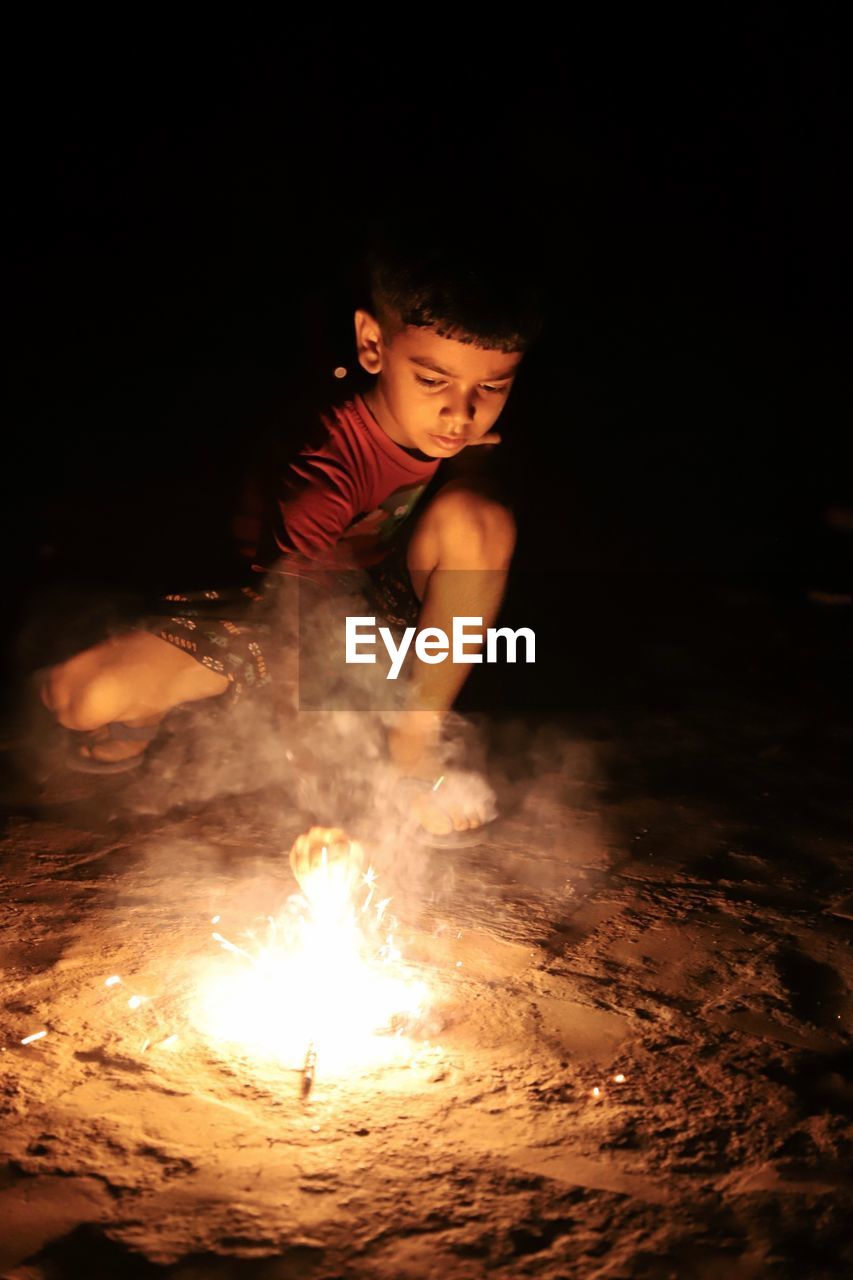 Cute boy crouching by fireworks on field at night