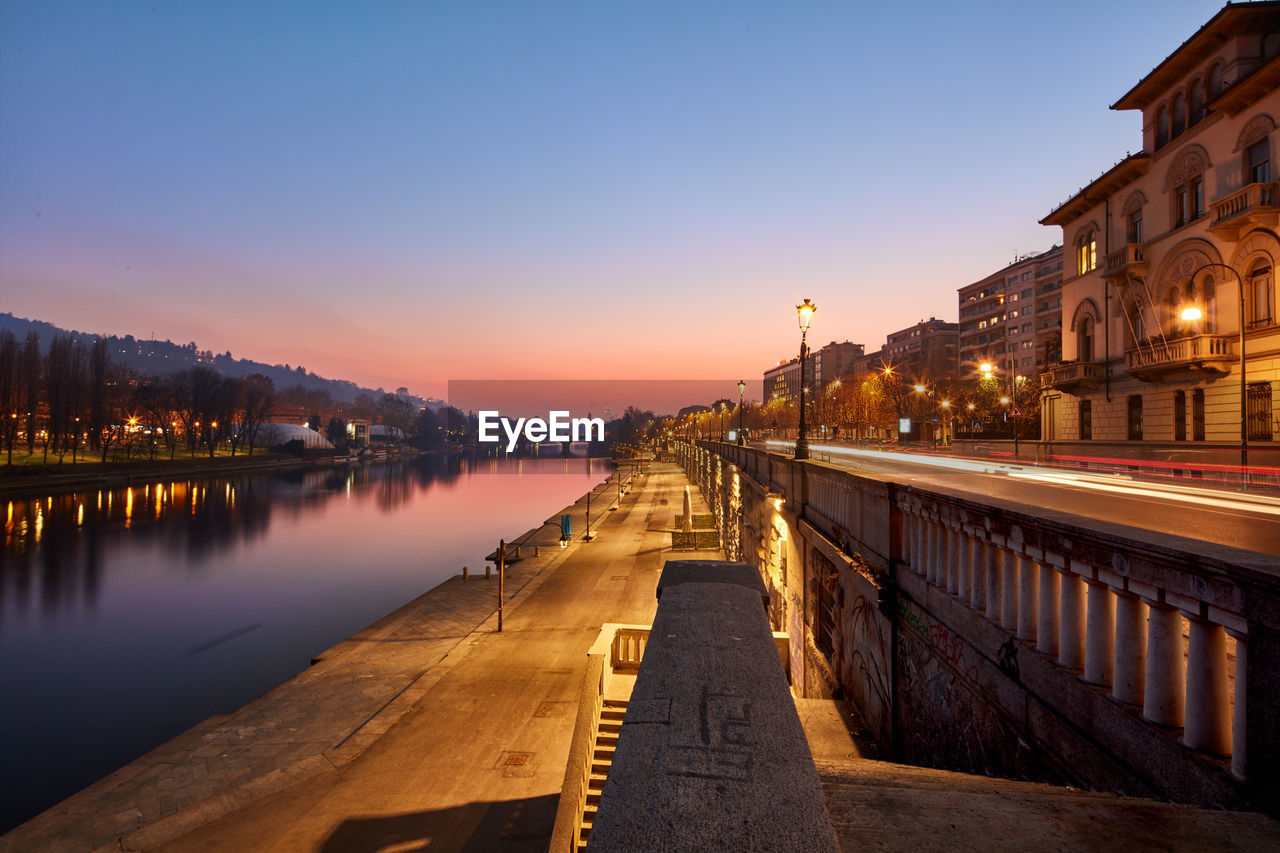 Illuminated buildings by river against sky at sunset