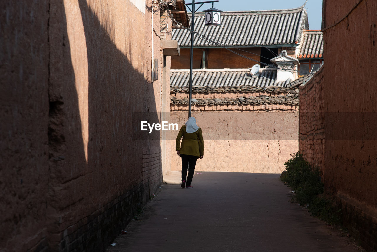 REAR VIEW OF MAN WALKING IN ALLEY AMIDST BUILDINGS IN CITY