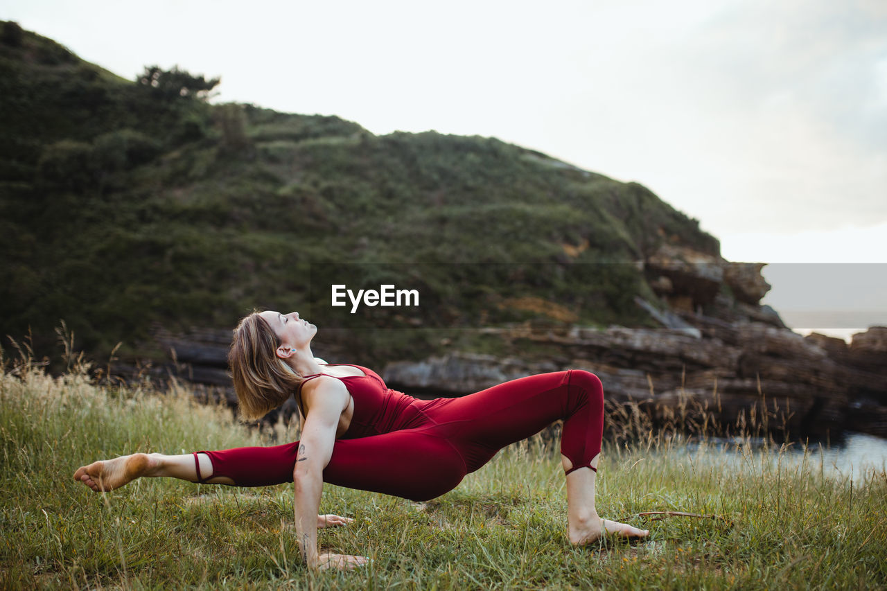 Young flexible woman in red sportswear standing in difficult yoga pose on empty green hill