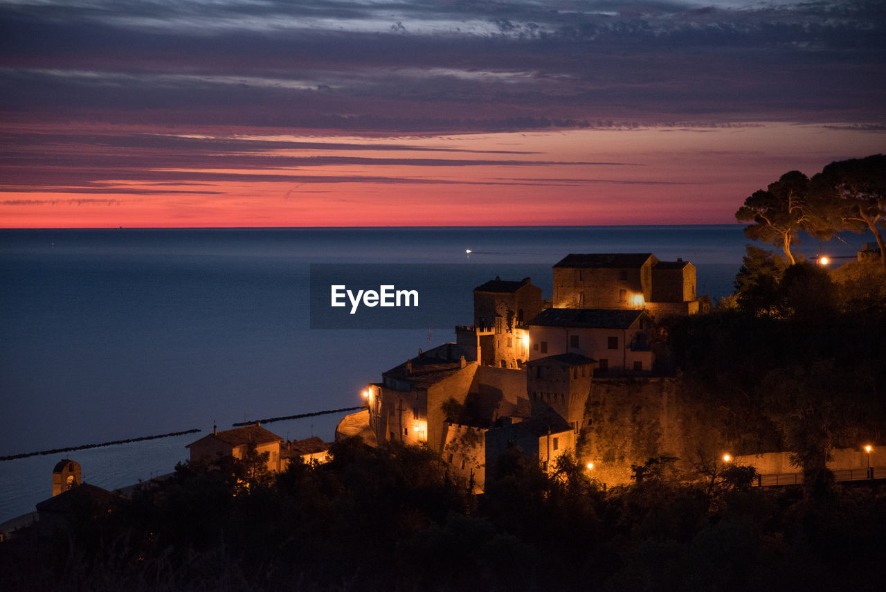 Illuminated historic village by sea against sky at sunset