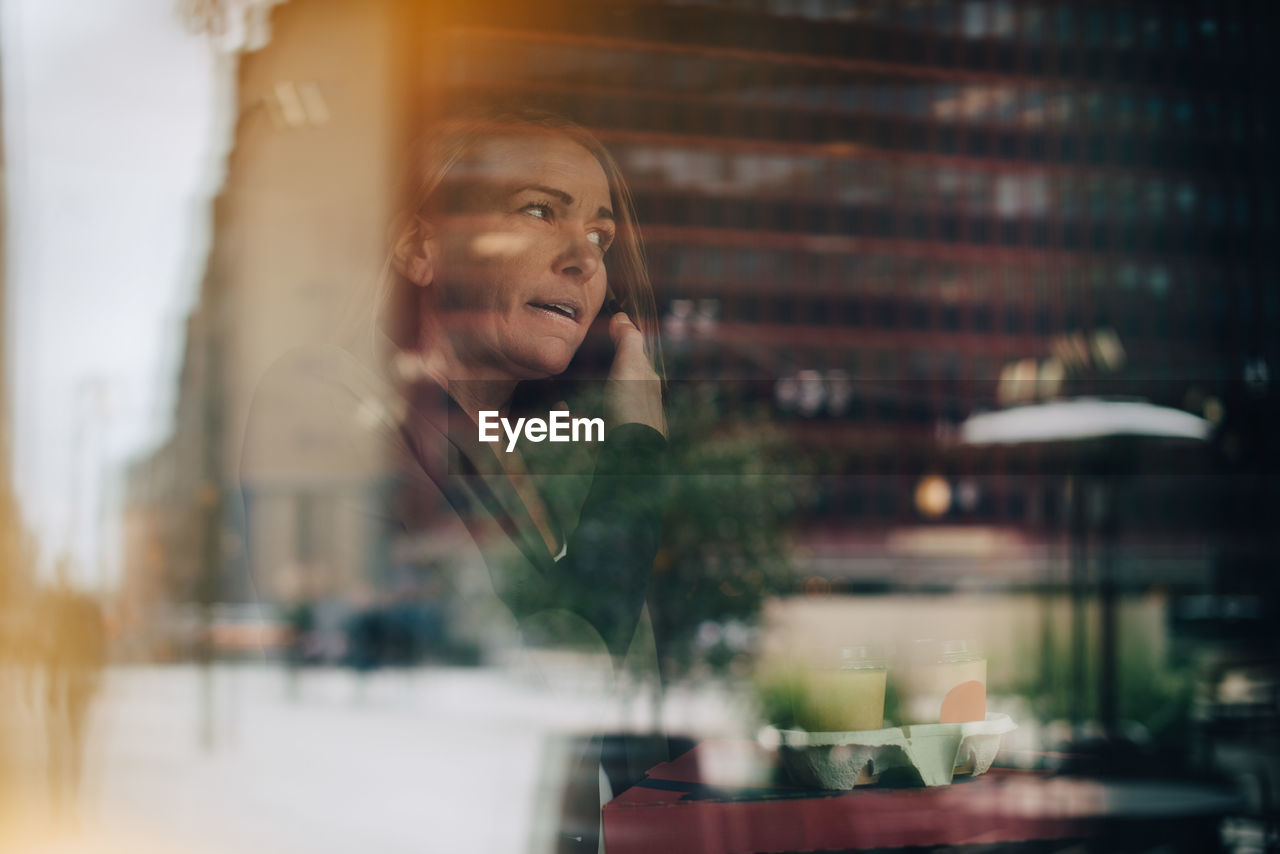 Mature businesswoman talking on mobile phone while holding food and drink in cafe seen from window