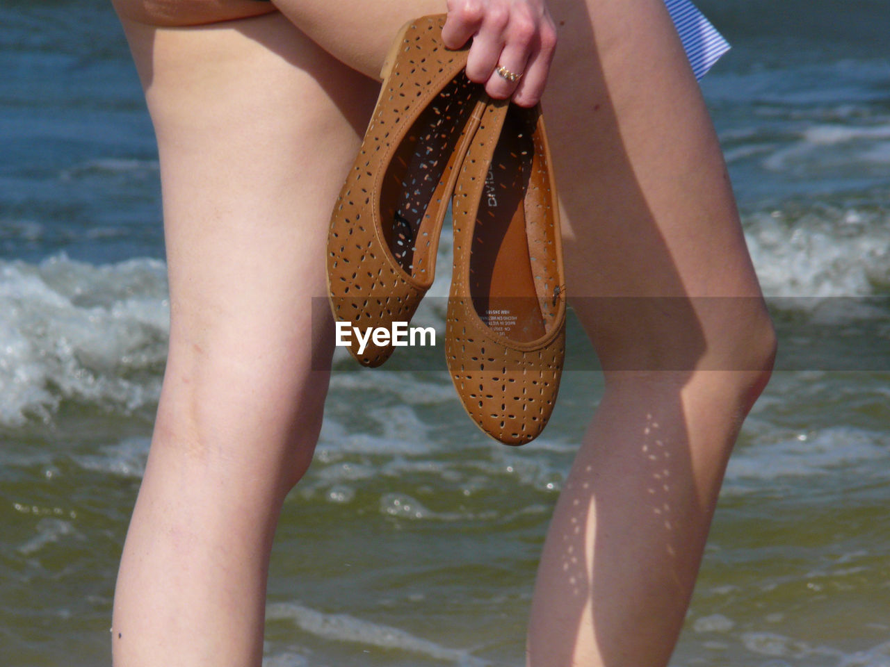 Midsection of woman holding shoes while walking at beach