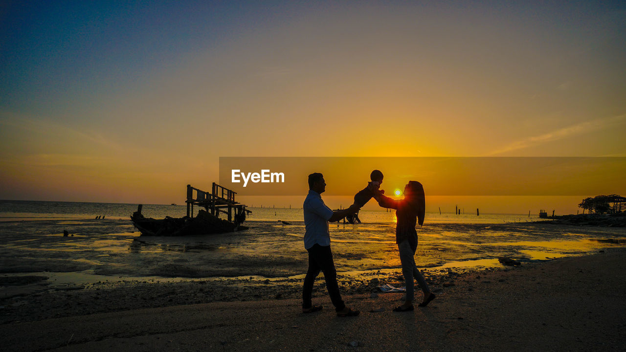 Parents holding baby while standing at beach against sky during sunset