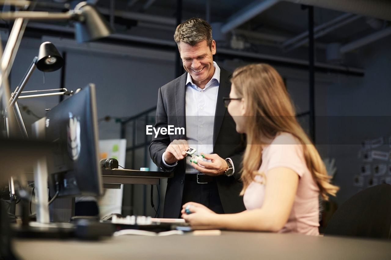 Creative businesswoman looking at smiling male bank manager holding solar toy car in creative office