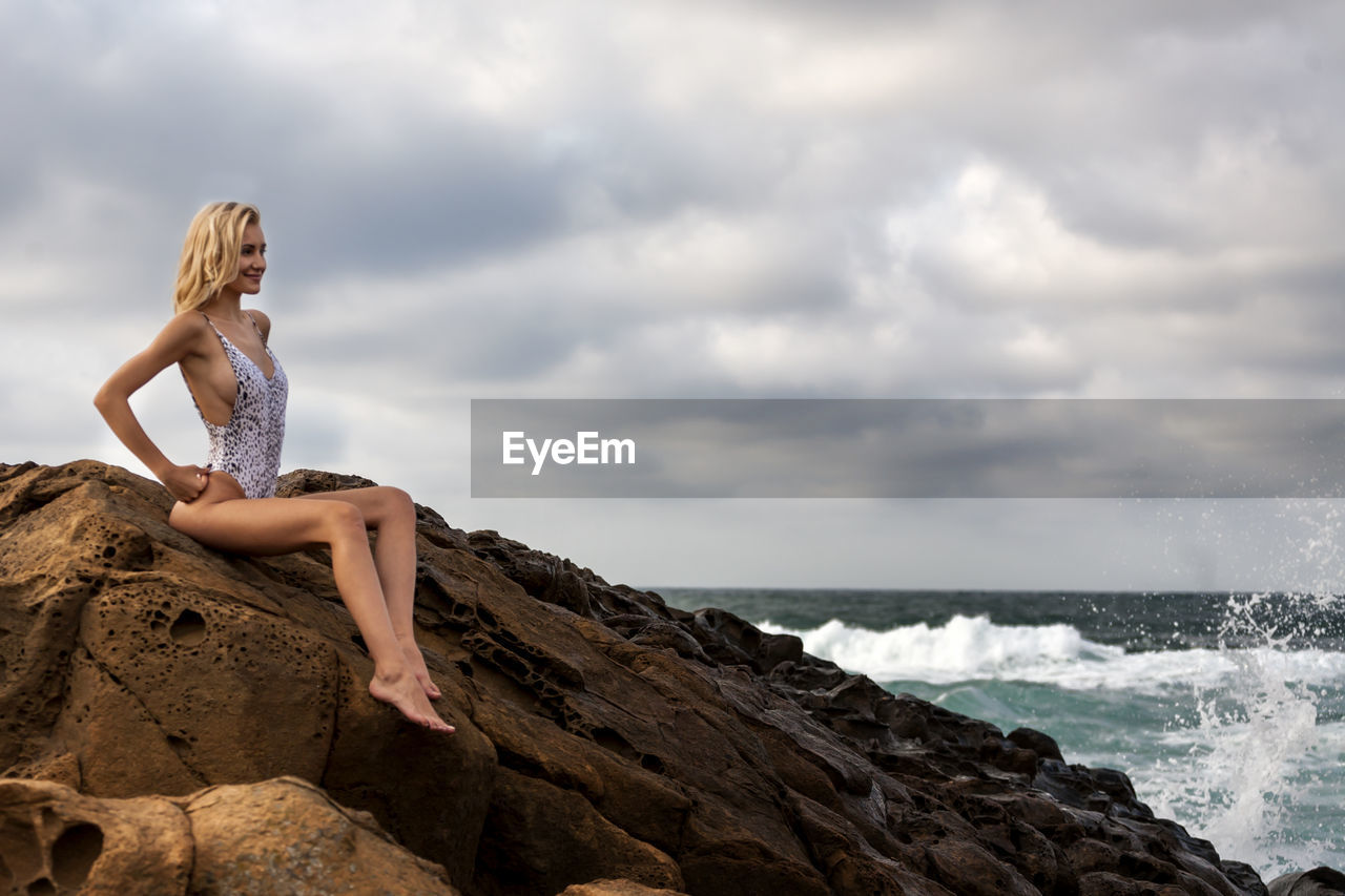 Side view of seductive woman looking at sea while sitting on rock against cloudy sky