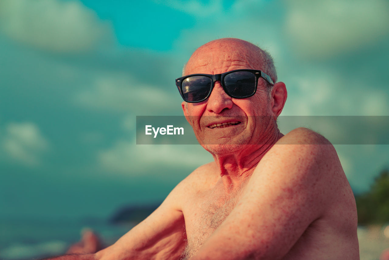 Portrait of shirtless man wearing sunglasses sitting at beach against sky