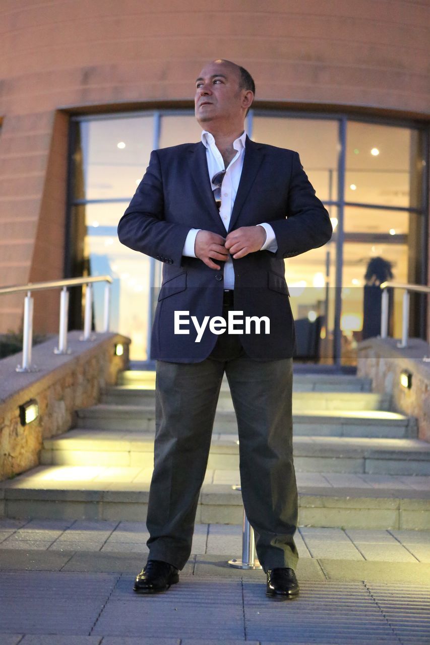 Businessman buttoning blazer while standing against building