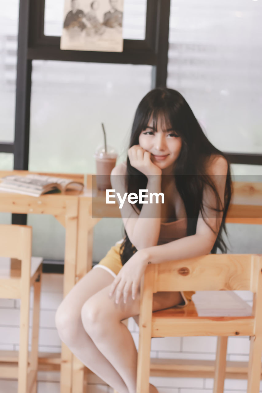 PORTRAIT OF YOUNG WOMAN SITTING ON CHAIR IN CAFE