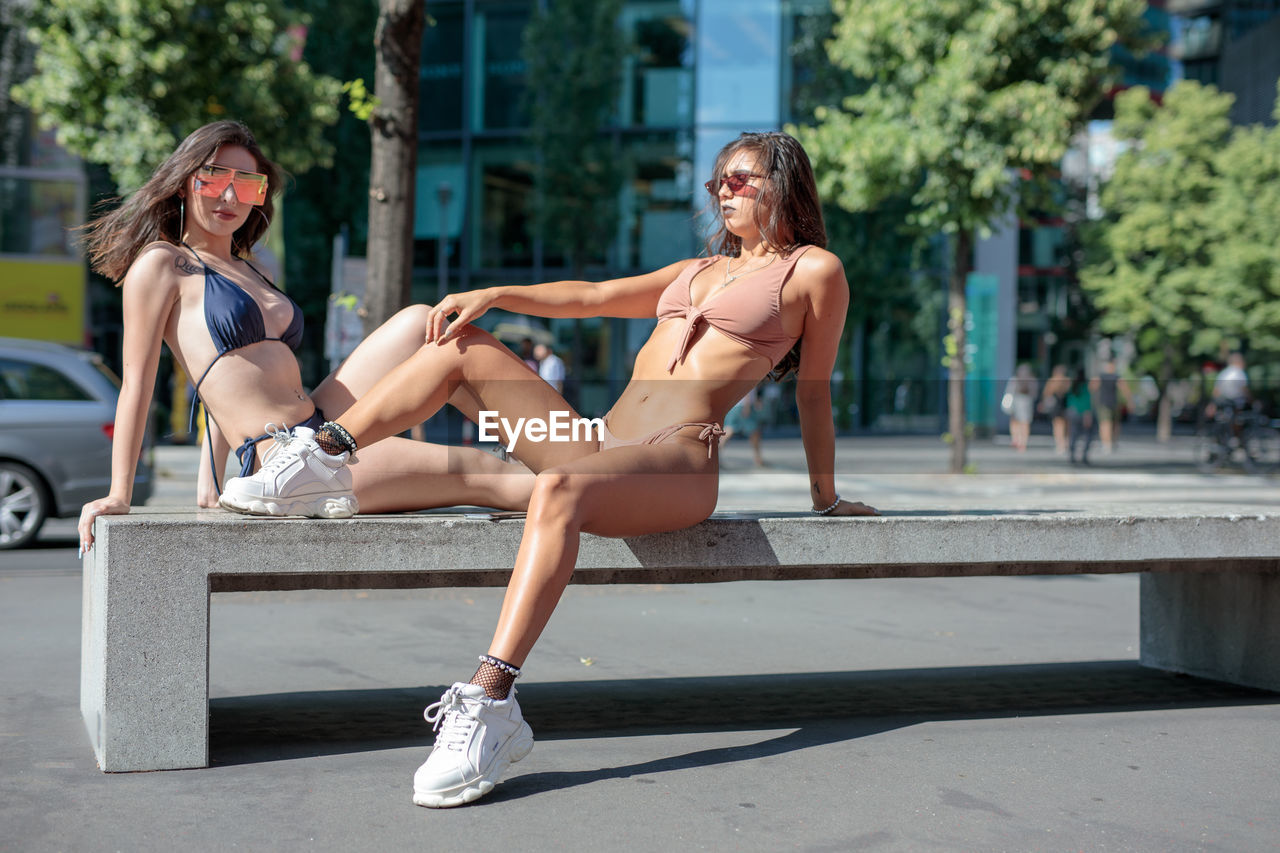Fashionable young women wearing bikinis while sitting in city during sunny day