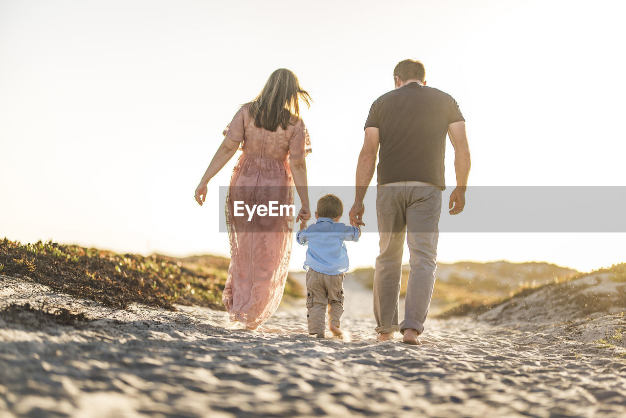 Rear view of parents holding son's hands while walking on sand at beach against clear sky during sunset