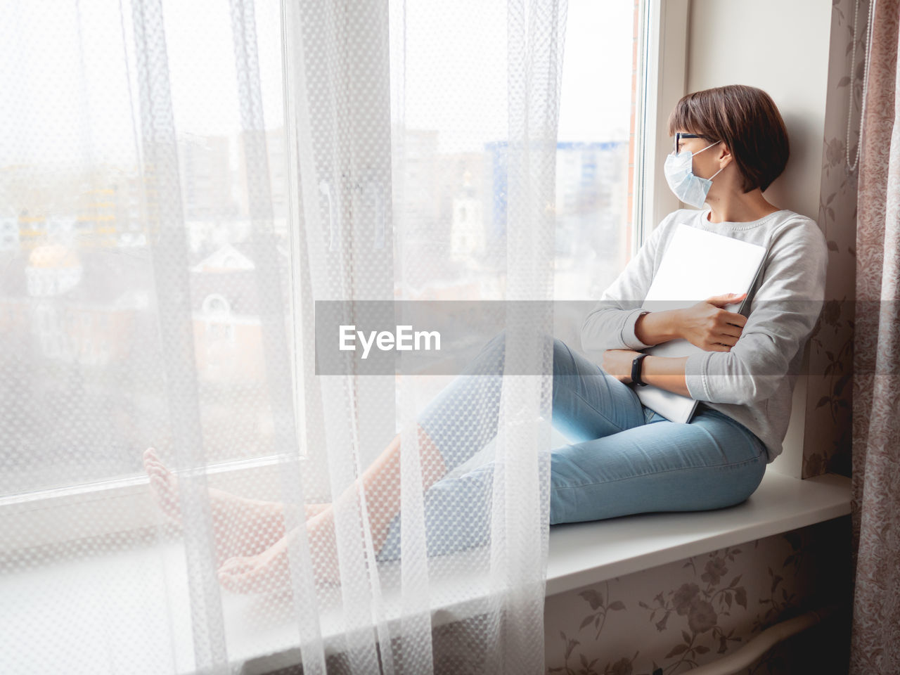Woman in medical mask had lost her job. she sits on window sill with closed laptop. 