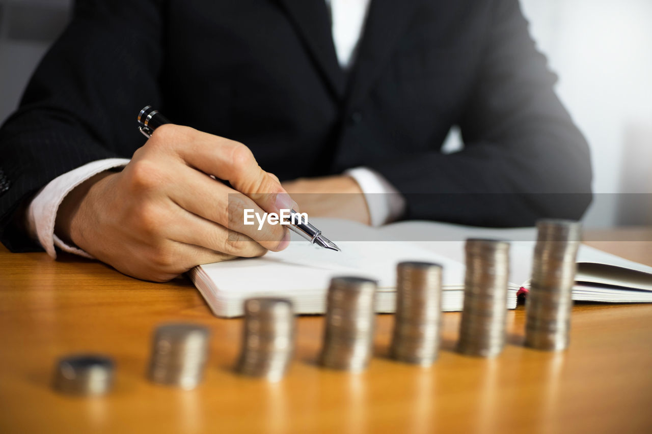 Midsection of man holding pen over book by coin stack on table