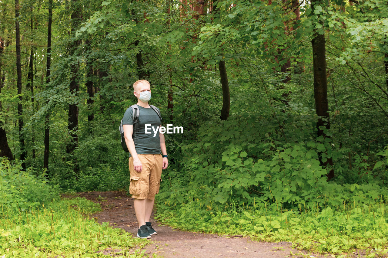 European young blond man in a disposable medical mask walks in the forest.