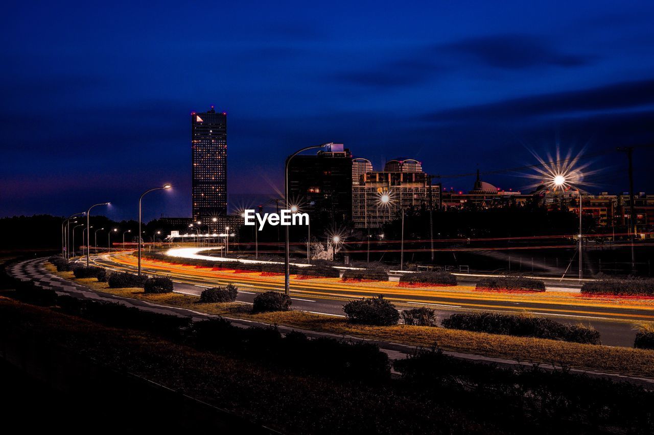 LIGHT TRAILS ON ROAD AGAINST ILLUMINATED BUILDINGS AT NIGHT