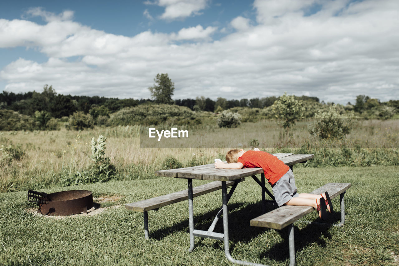 Side view of boy lying on picnic bench against cloudy sky during sunny day