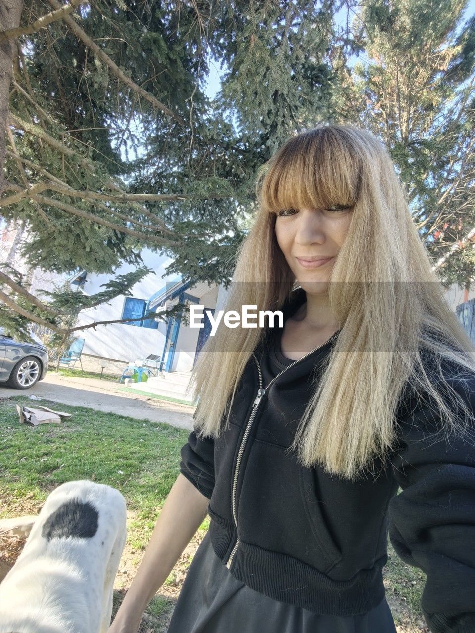 one person, tree, long hair, blond hair, women, leisure activity, hairstyle, plant, clothing, young adult, adult, day, lifestyles, standing, casual clothing, nature, spring, three quarter length, human hair, person, portrait, smiling, front view, waist up, outdoors, dog, motor vehicle, transportation, car, mode of transportation, looking at camera, fashion, emotion, looking, happiness