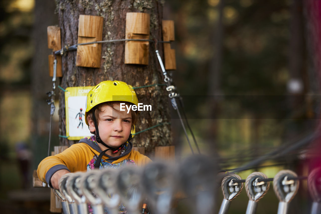 Boy in yellow helmet standing on suspension bridge near tree trunk on summer weekend day in adventure park in forest