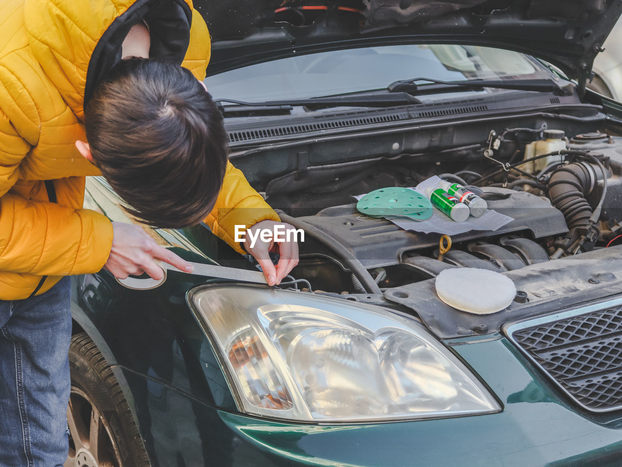 A guy in a yellow jacket sticks scoich around the edges of the headlight preparing it for polishing