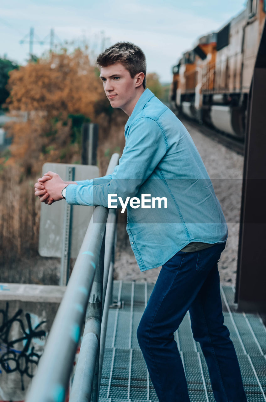 Side view of young man standing by railing against railroad track