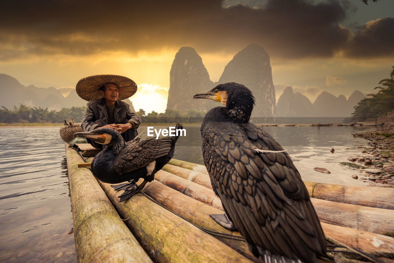 Man wearing hat sitting on raft with bird against mountains and sky