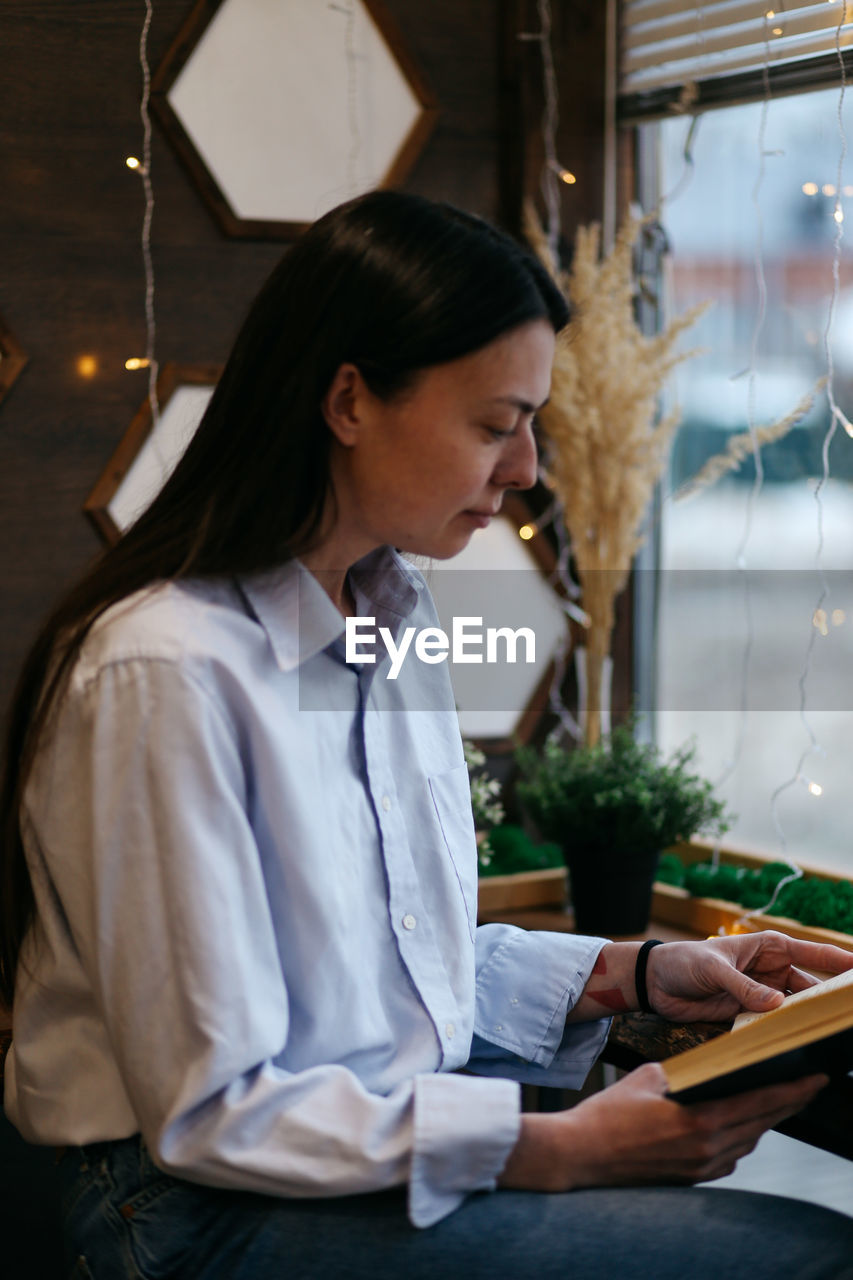 Brunette girl by the window in a cafe drinking coffee, reading a book
