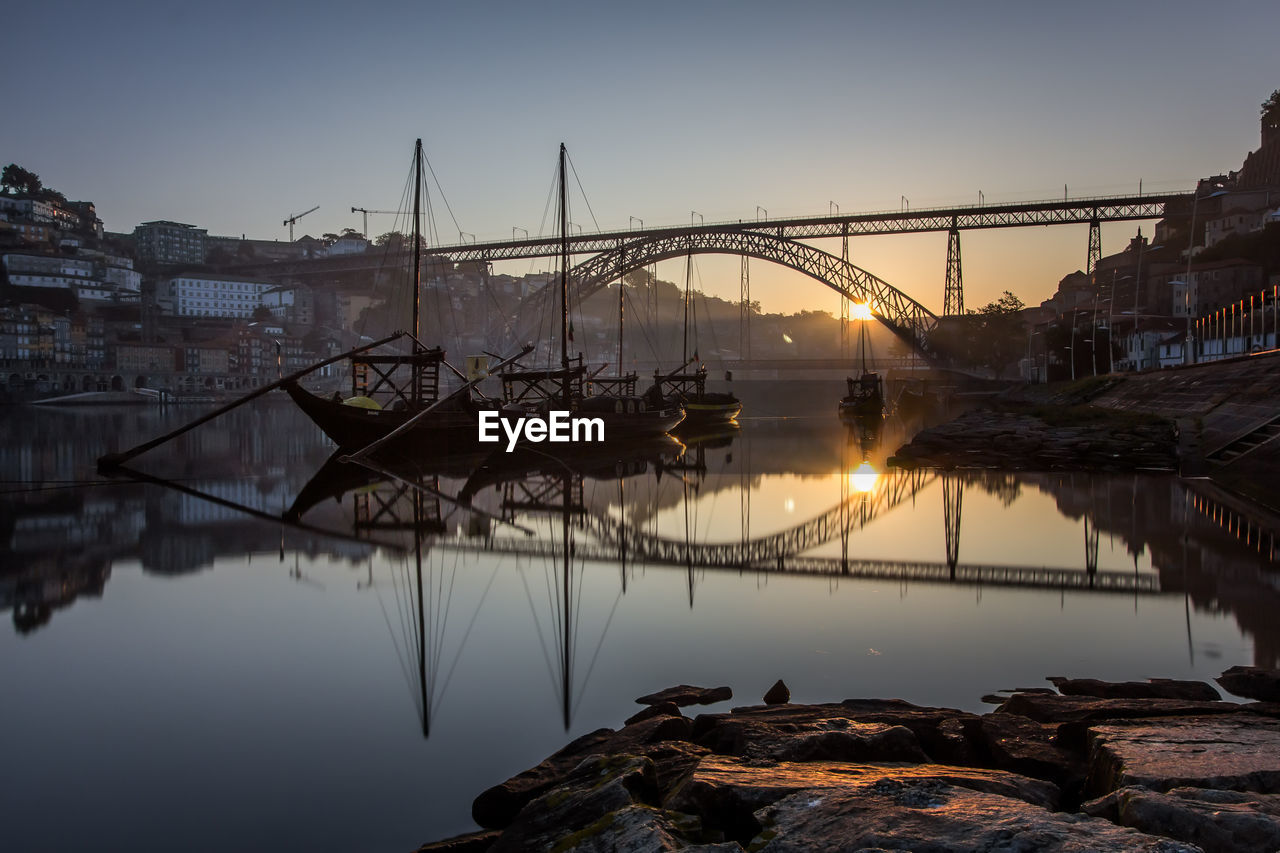 Reflection of bridge by boats moored on river at sunset