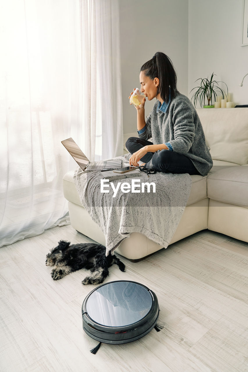 Woman sitting on sofa in light room working on a computer and drinking cold beverage with cute puppy lying down next round black robotic vacuum cleaner