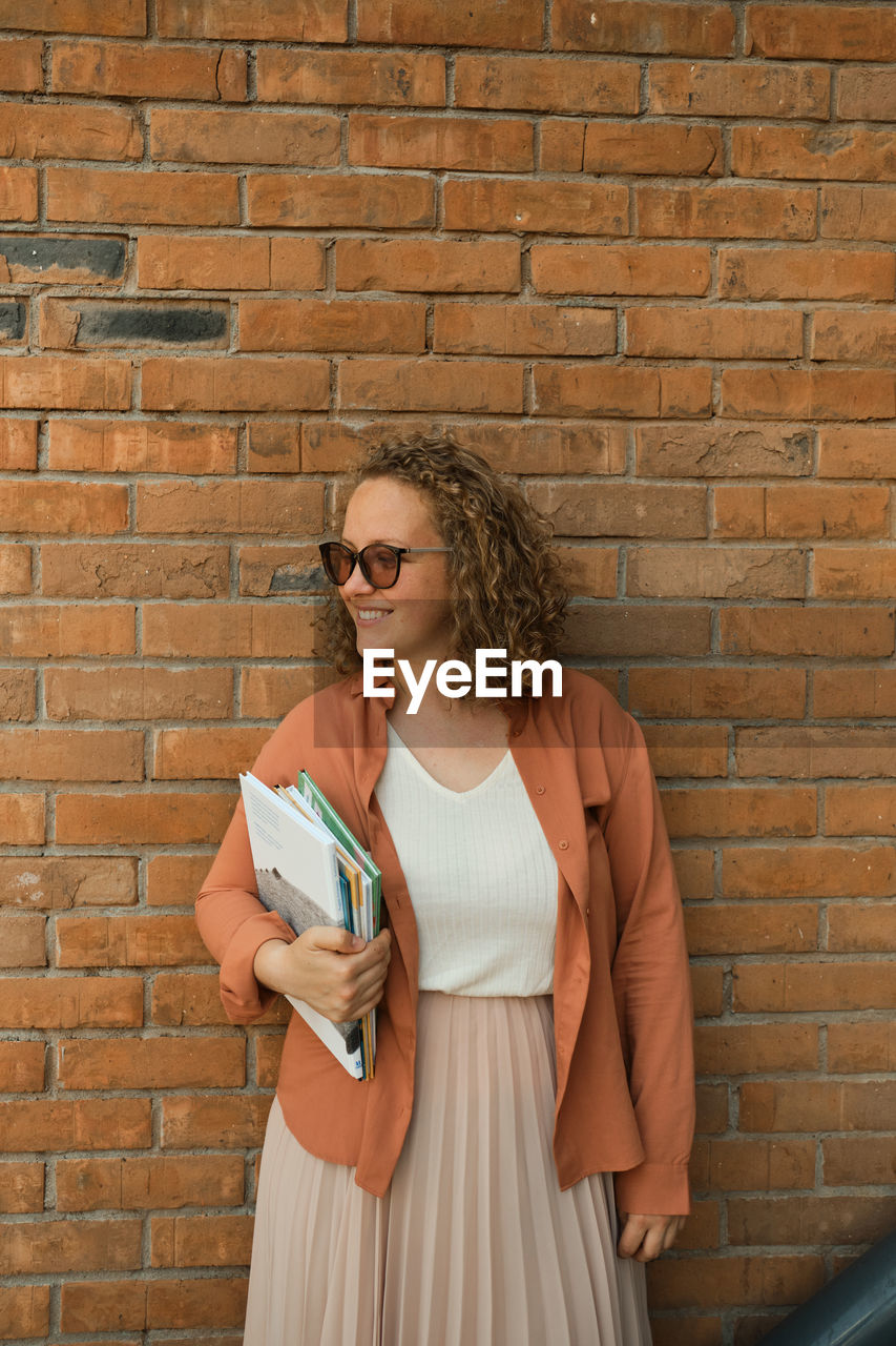 Young adult millennial with curly hair smiles. woma glasses. student holds book. lifestyle in city