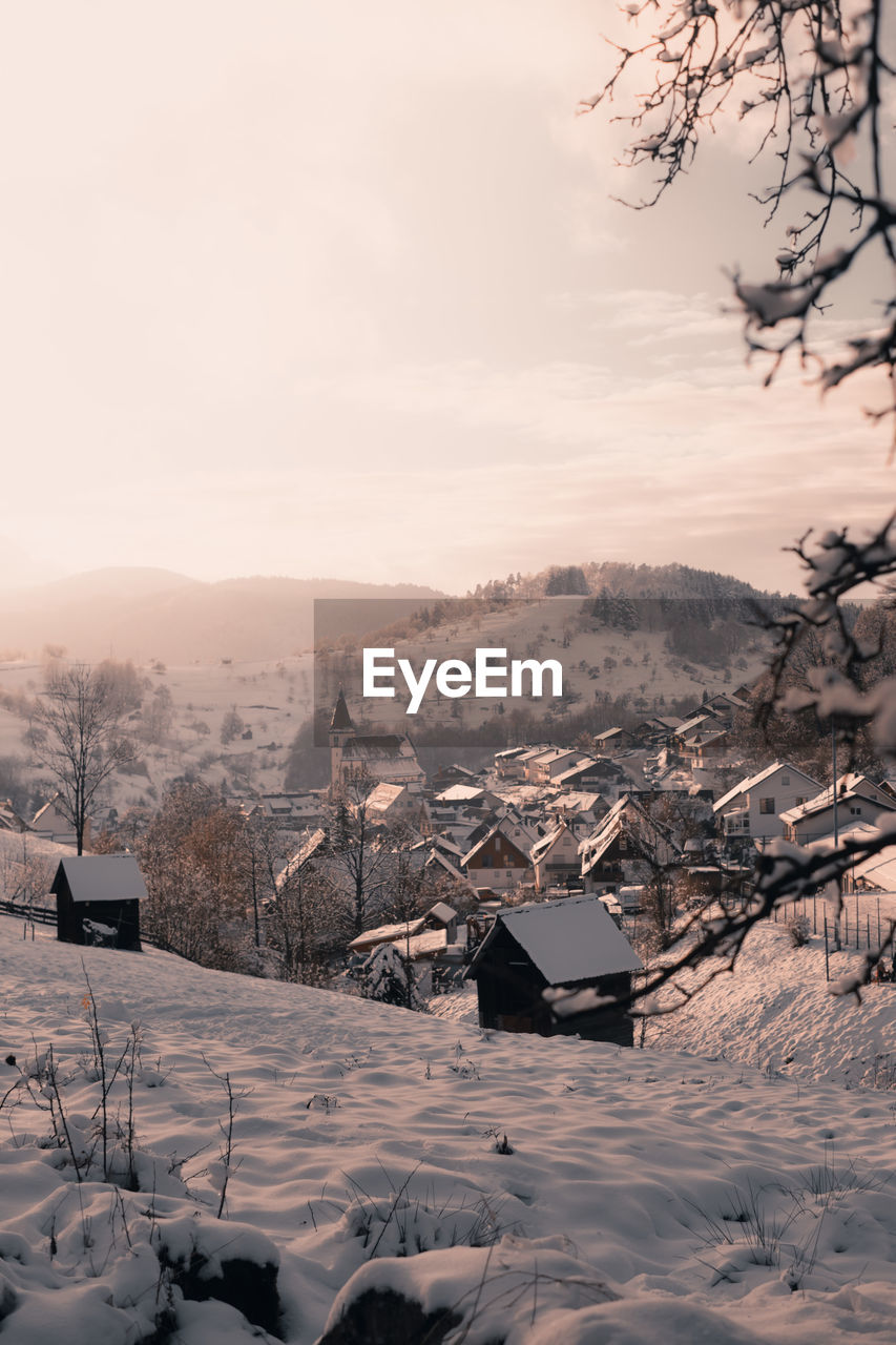 Snow covered houses by buildings against sky and mountains during winter. branches in foreground.