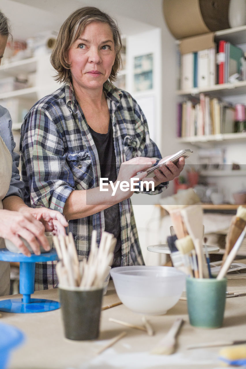 Mature female potter using mobile phone by senior colleague molding clay at workbench in store