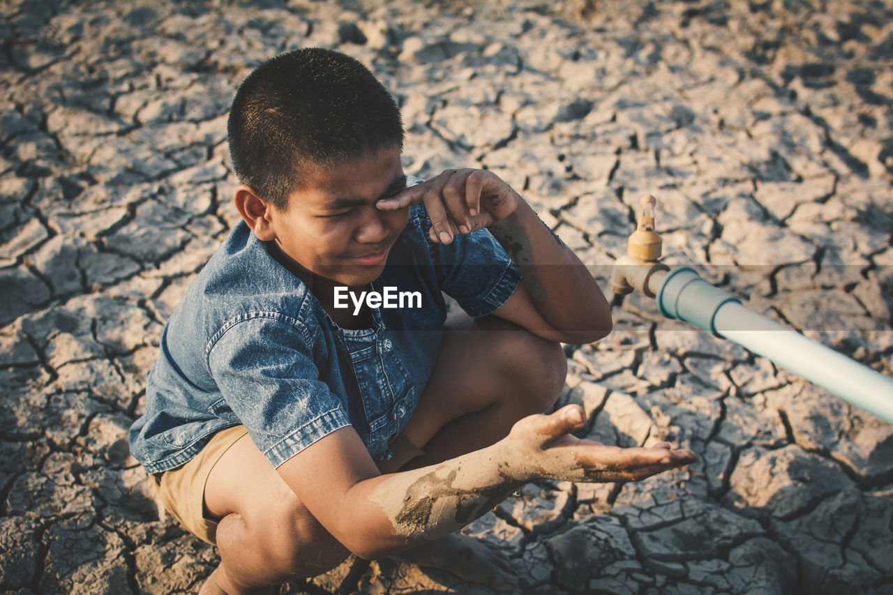 Boy crying by faucet on cracked field