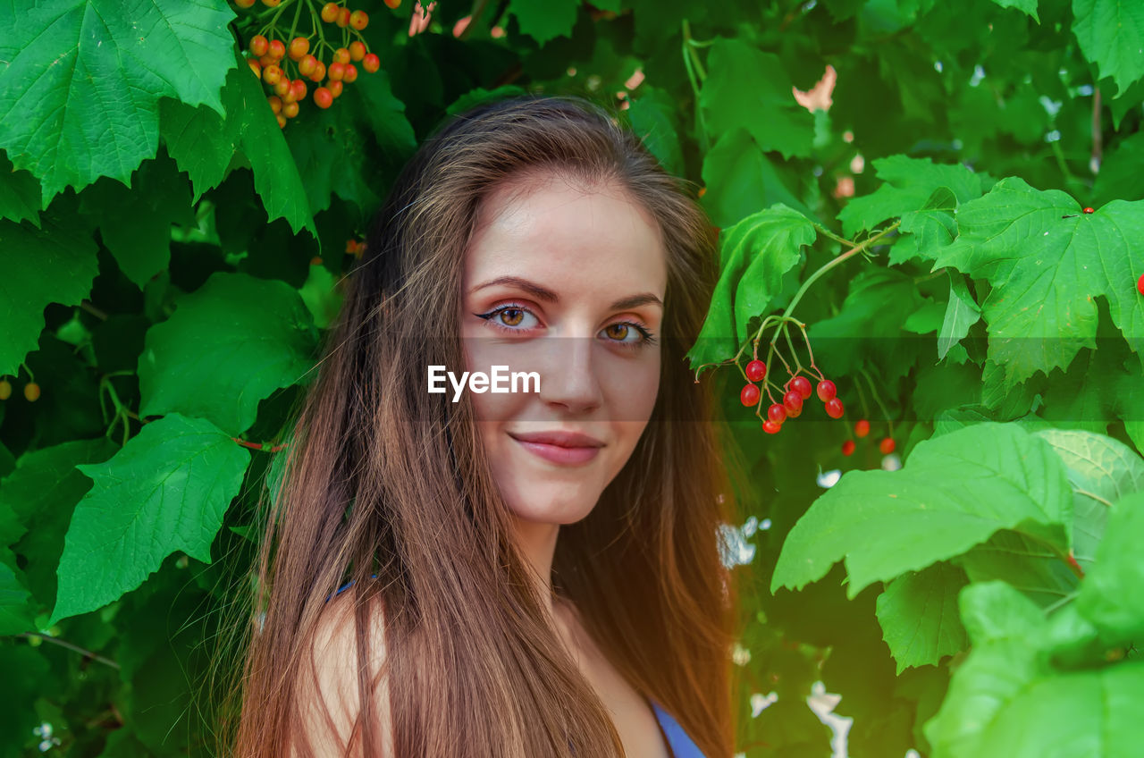 Attractive woman with long hair looks into the camera with half turn on background of viburnum bush