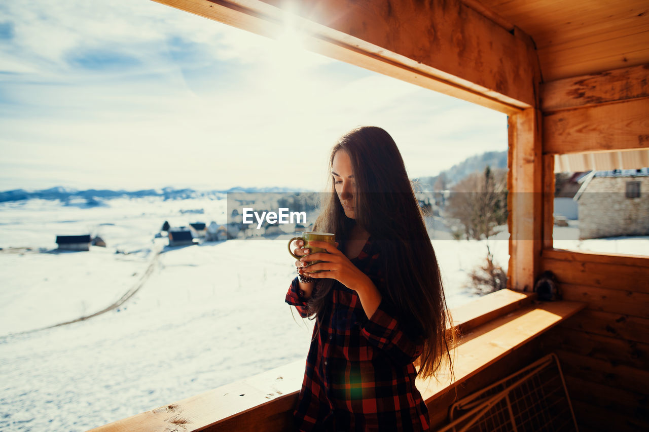 Portrait of young woman holding cup while standing by window in cottage during winter