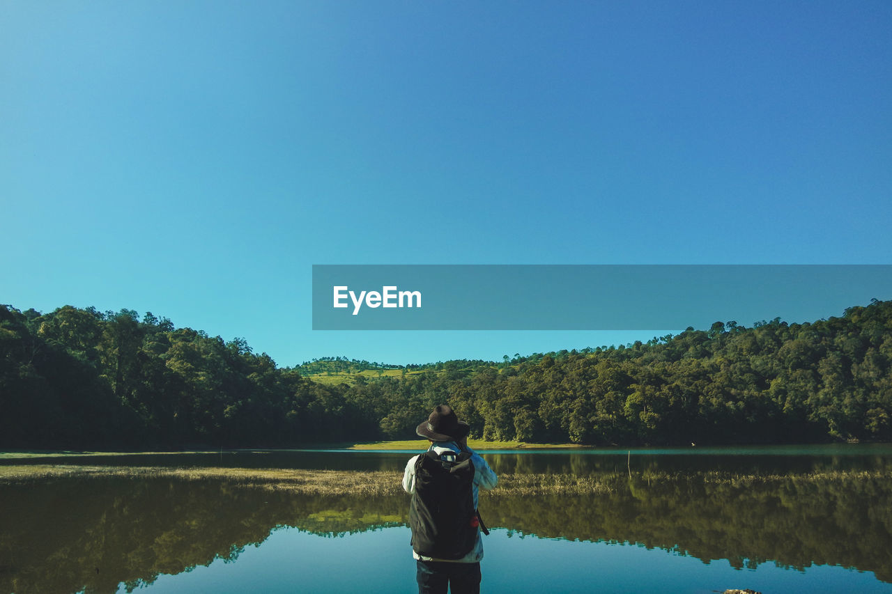 Rear view of man looking at lake against clear blue sky